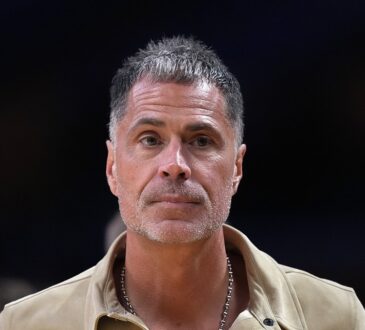 Los Angeles Lakers general manager Rob Pelinka, wearing a tan suede shirt jacket, looks on from courtside.