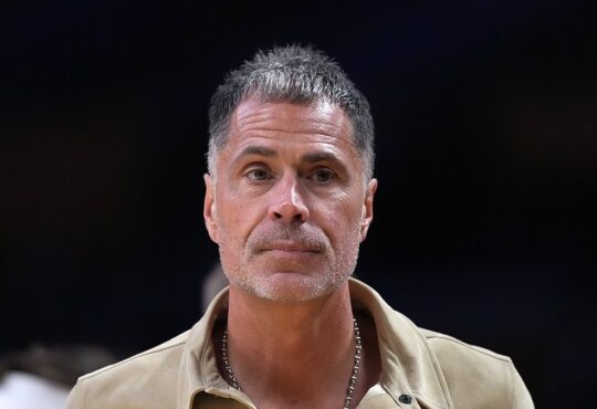 Los Angeles Lakers general manager Rob Pelinka, wearing a tan suede shirt jacket, looks on from courtside.