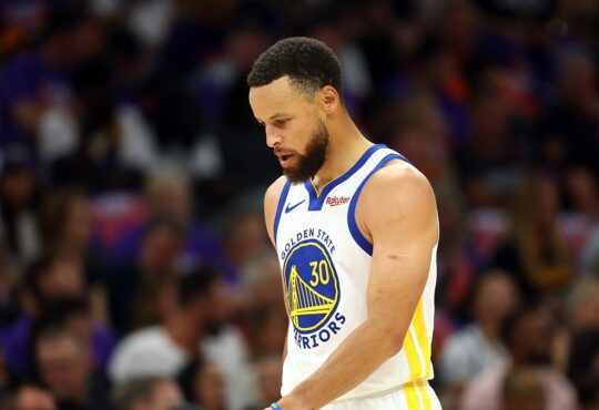 Golden State Warriors guard Stephen Curry looks down as he walks across the court in a white Warriors jersey.